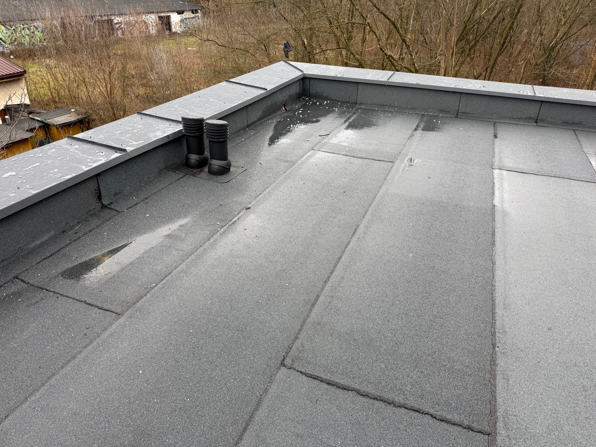 Widok z góry na płaski dach pokryty papą termozgrzewalną z dwoma kominkami wentylacyjnymi w Legionowie, widoczne drobne kałuże po deszczu i obróbki blacharskie na krawędzi dachu.