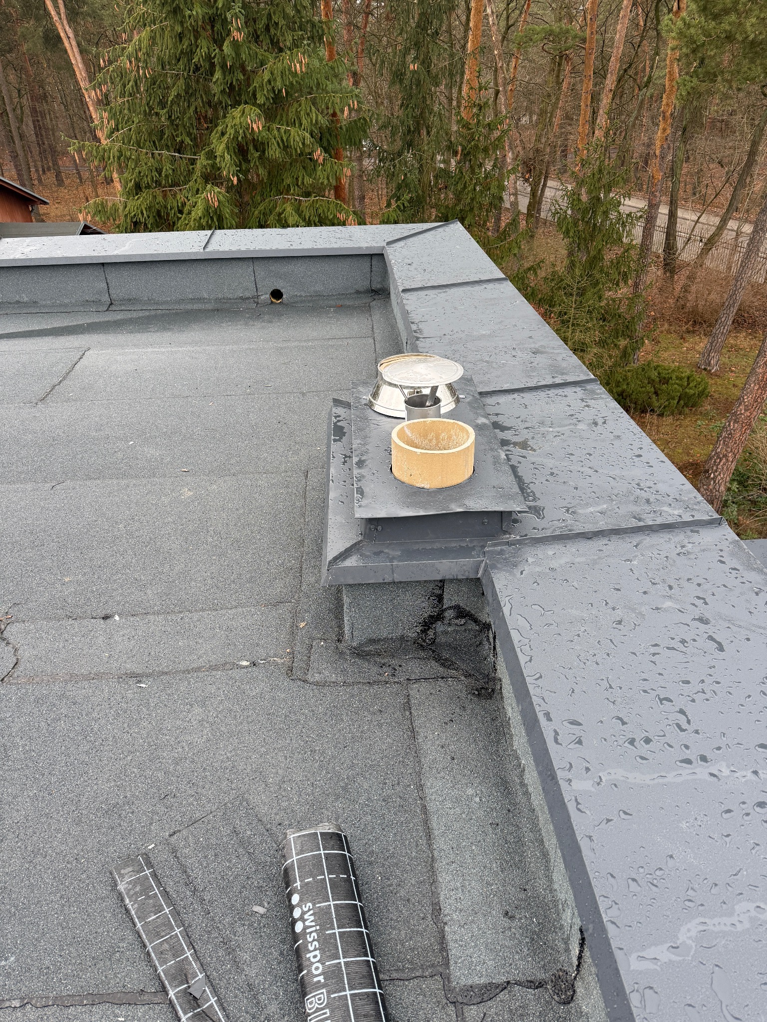 Szary, płaski dach pokryty papą, z widocznymi detalami wykończeniowymi, takimi jak komin wentylacyjny i obróbki blacharskie, oraz rolka papy na powierzchni dachu.