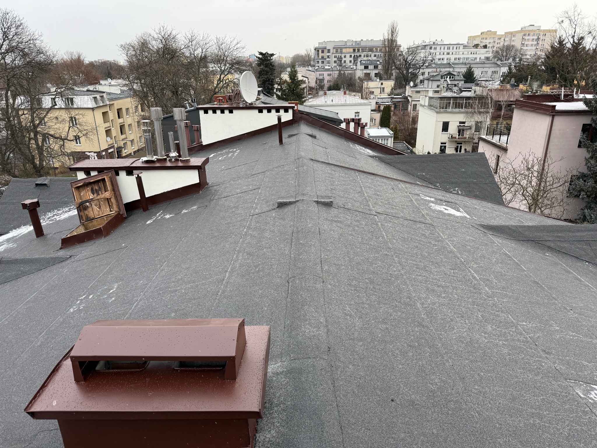 Bitumiczna powierzchnia dachu z widocznymi kominami i anteną satelitarną na tle zabudowy miejskiej w pochmurny dzień.