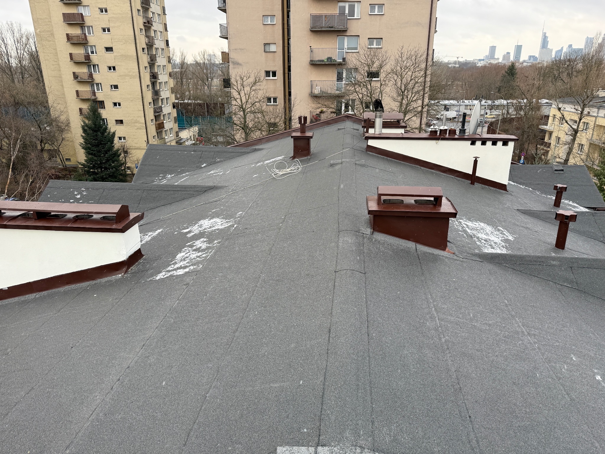 Widok z góry na płaski dach pokryty papą, z widocznymi kominami w brązowo-białej kolorystyce, delikatnie przyprószony śniegiem, z budynkami mieszkalnymi i panoramą miasta w tle.