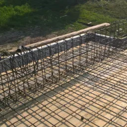 Siatka zbrojeniowa ułożona na deskowaniu fundamentu, widoczny fragment zbrojenia pionowego, w tle zieleń, obok leży szlifierka kątowa na małym stoliku.