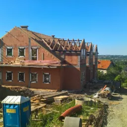 Dwukondygnacyjny budynek w trakcie budowy z czerwonej cegły, widoczne drewniane elementy konstrukcji dachu, okna wstawione, na pierwszym planie sterta ziemi i przenośna toaleta.