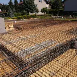 Zbrojenie płyty fundamentowej na styropianie, widoczny szalunek obwodowy i fragmenty muru z bloczków betonowych w tle.