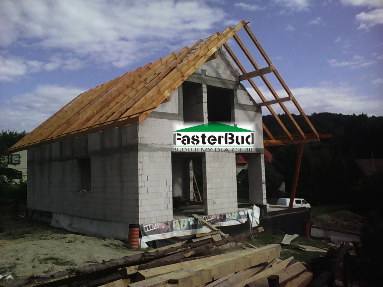Surowy stan domu z pustaków z drewnianą konstrukcją dachu w trakcie budowy, widoczny fundament i materiały budowlane na pierwszym planie, logo firmy nałożone na zdjęcie.