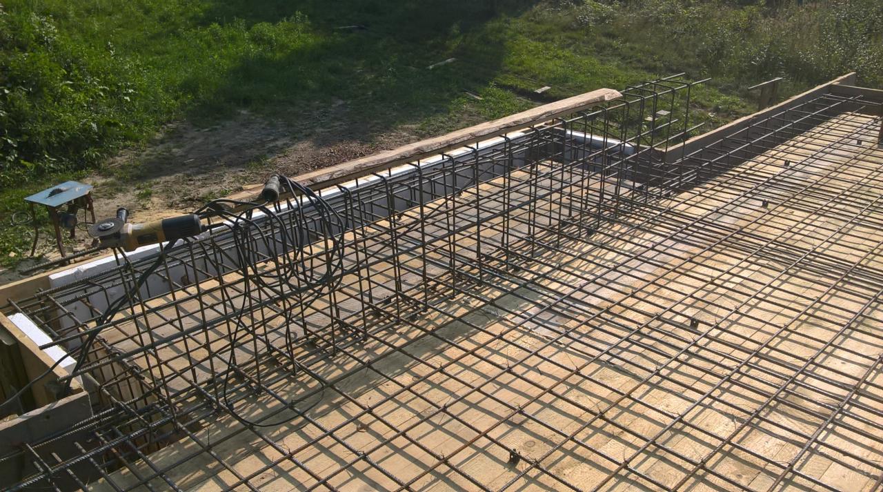 Siatka zbrojeniowa ułożona na deskowaniu fundamentu, widoczny fragment zbrojenia pionowego, w tle zieleń, obok leży szlifierka kątowa na małym stoliku.