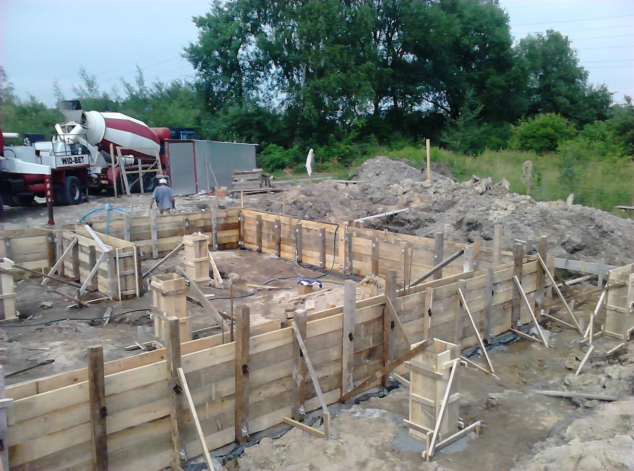 Deskowanie fundamentów pod budynek z betoniarką Wid-Bet w tle, widoczne drewniane szalunki i przygotowany teren budowy. Prace fundamentowe w toku.