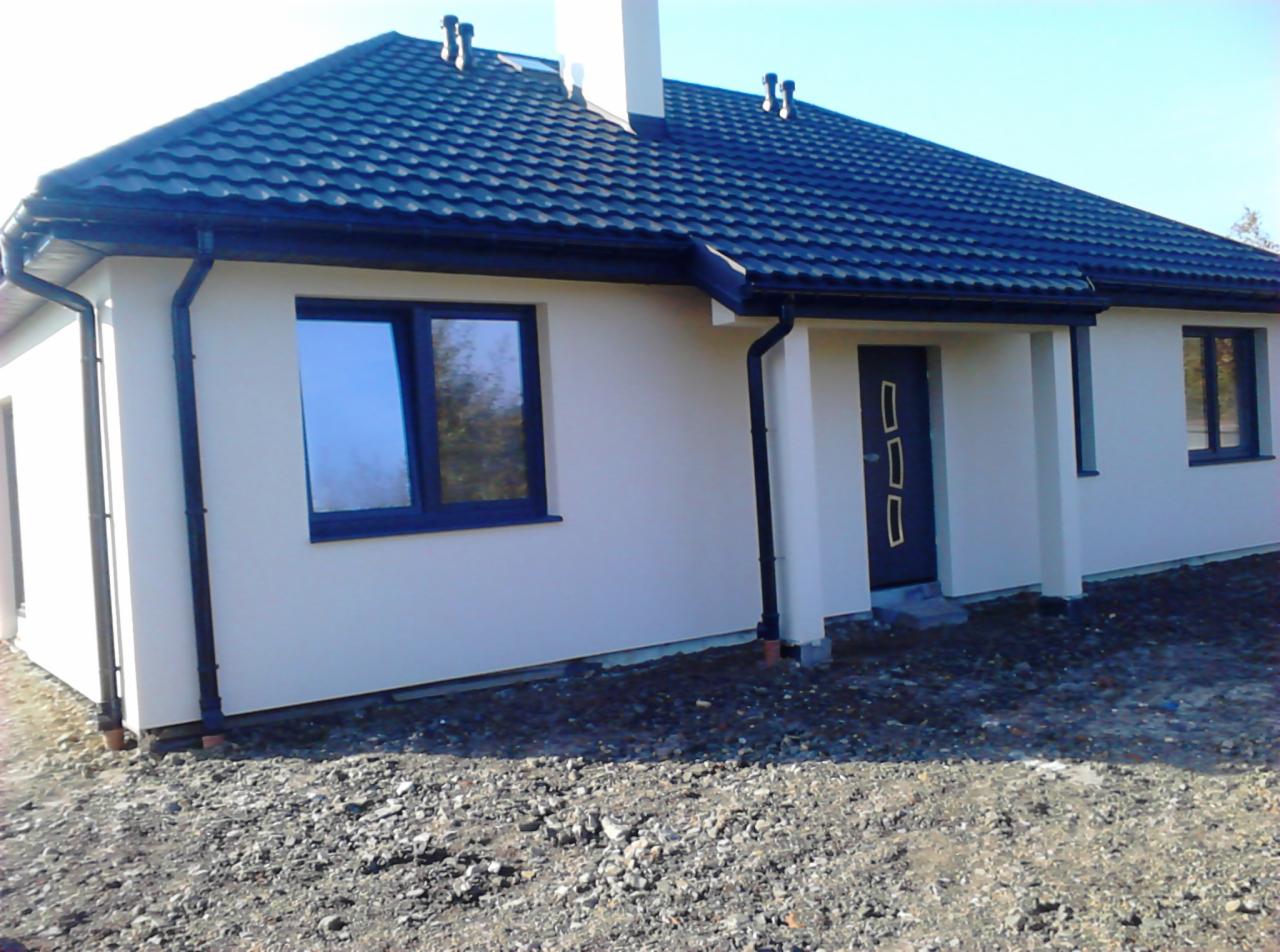 Gotowy dom z ciemną dachówką i stolarką okienną, elewacja w jasnym odcieniu, widoczne rynny i fragment niezagospodarowanego terenu przed budynkiem.