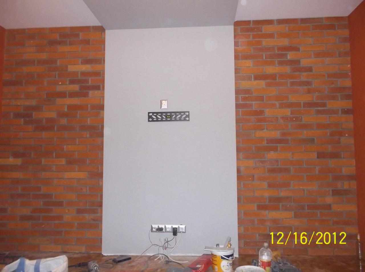 Pokój w trakcie prac wykończeniowych: fragment ściany z cegłą, szara ściana z gniazdkami i uchwytem na TV, farba i narzędzia na podłodze. Data na zdjęciu: 16.12.2012.