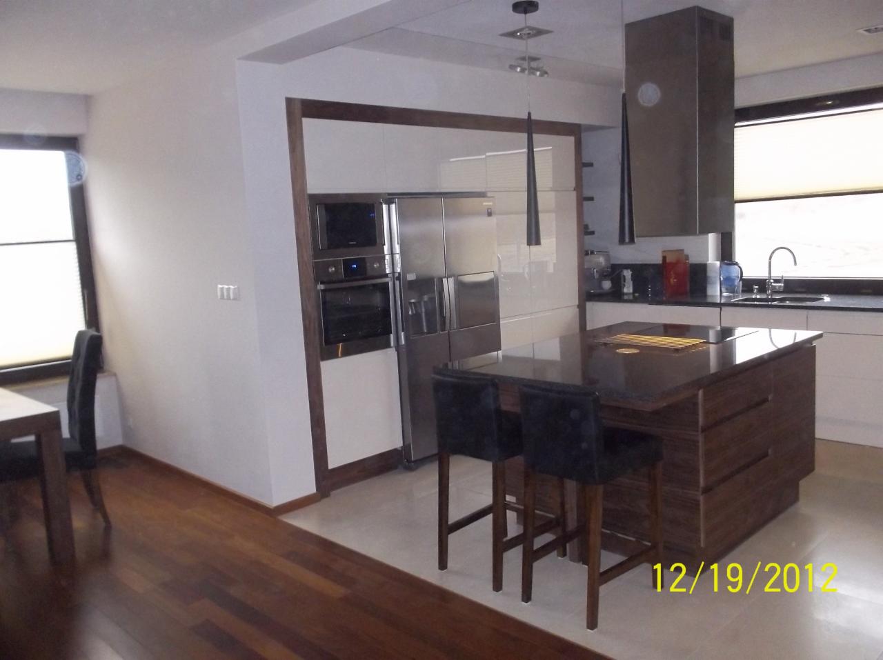 Nowoczesna kuchnia z wyspą i drewnianą podłogą. Zabudowa kuchenna z białymi frontami i drewnianym obramowaniem. Stalowy sprzęt AGD.