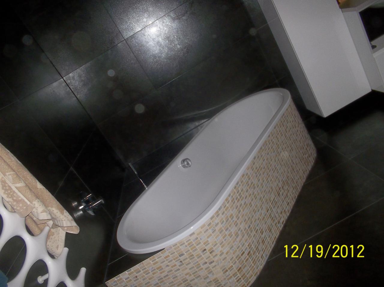 Nowoczesna wanna obudowana mozaiką w wykończonej łazience z ciemną, elegancką podłogą. Widoczny fragment szafki i wieszak na ręczniki.