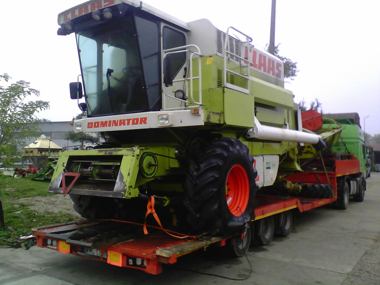 Kombajn zbożowy Claas Dominator na czerwonej platformie niskopodwoziowej z pomarańczowymi pasami mocującymi, widok z tyłu, tablica rejestracyjna widoczna.