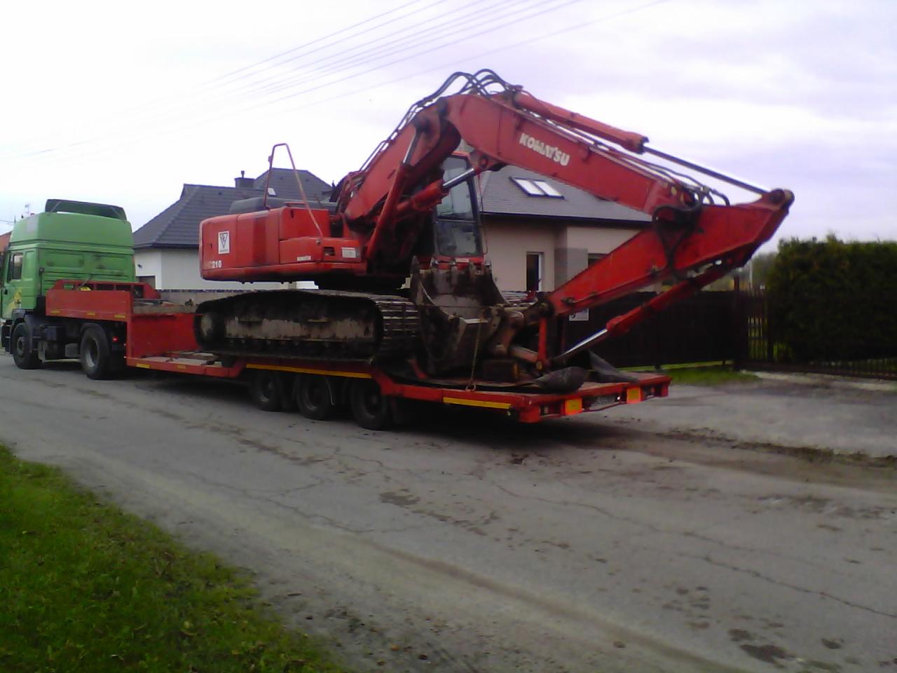 Czerwona koparka Komatsu załadowana na niskopodwoziową platformę, ciągniętą przez zielony ciągnik siodłowy, na drodze w Markach.