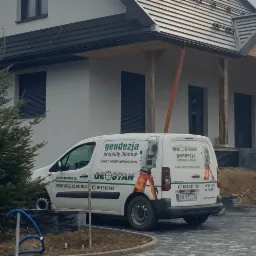 Nowoczesny dom jednorodzinny z szarą dachówką, na posesji stoi biały samochód dostawczy z logo firmy geodezyjnej Geostan oraz szary samochód osobowy Suzuki. Widoczne drzewa iglaste i ułożona kostka...