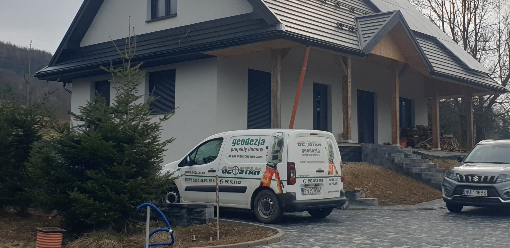 Nowoczesny dom jednorodzinny z szarą dachówką, na posesji stoi biały samochód dostawczy z logo firmy geodezyjnej Geostan oraz szary samochód osobowy Suzuki. Widoczne drzewa iglaste i ułożona kostka...
