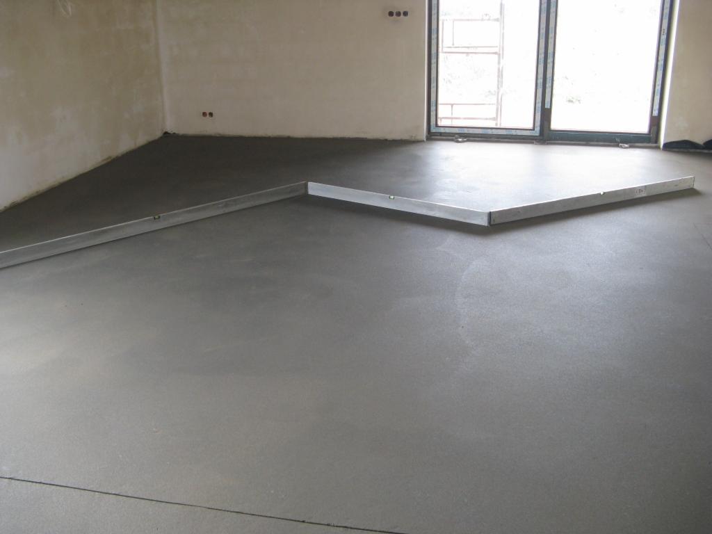 Świeżo wylana, szara posadzka z widocznymi łatami poziomującymi w jasnym pomieszczeniu z oknami.