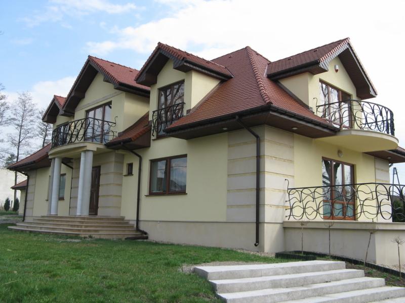 Dwukondygnacyjny dom jednorodzinny z beżową elewacją, balkonami z kutymi balustradami i schodami wejściowymi wyłożonymi płytami w jasnym odcieniu szarości.