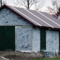 Surowy budynek gospodarczy z szarego bloczka betonowego z ciemnozielonymi przesuwnymi wrotami, z brązowym dachem z trzema jasnymi pasami, na tle wiejskiej zabudowy i silosów.