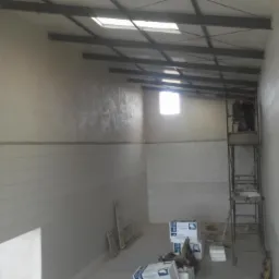 Wnętrze hali w trakcie wykończenia, widoczne ściany z płyt warstwowych, stalowa konstrukcja dachu i pracownicy na rusztowaniu. Częściowo otynkowana ściana.