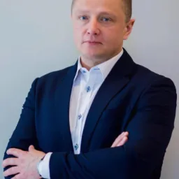 Portret mężczyzny w granatowym garniturze i białej koszuli, z założonymi rękami, patrzącego prosto w obiektyw na tle jasnoszarej ściany. Widoczny zegarek na lewym nadgarstku.