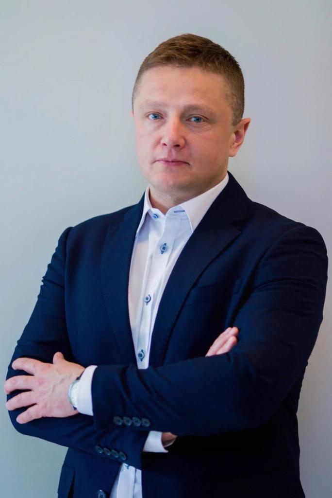 Portret mężczyzny w granatowym garniturze i białej koszuli, z założonymi rękami, patrzącego prosto w obiektyw na tle jasnoszarej ściany. Widoczny zegarek na lewym nadgarstku.