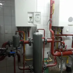 Dwa białe kotły grzewcze wiszące na ścianie z białych płytek, połączone systemem czerwonych i żółtych rur z manometrami i zaworami, widoczne szare zbiorniki wyrównawcze.