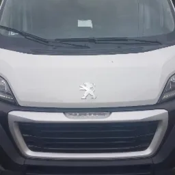Przód białego samochodu dostawczego Peugeot z widocznym logo na masce, reflektorami i czarnym zderzakiem. Lekko pochmurne tło.