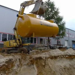 Żółty zbiornik z logo Hauraton podnoszony przez koparkę na tle budynku przemysłowego i wykopu w ziemi, osoba filmuje akcję telefonem.