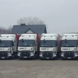 Flota białych ciężarówek z logo firmy transportowej zaparkowana na placu, widok z przodu, pochmurny dzień.