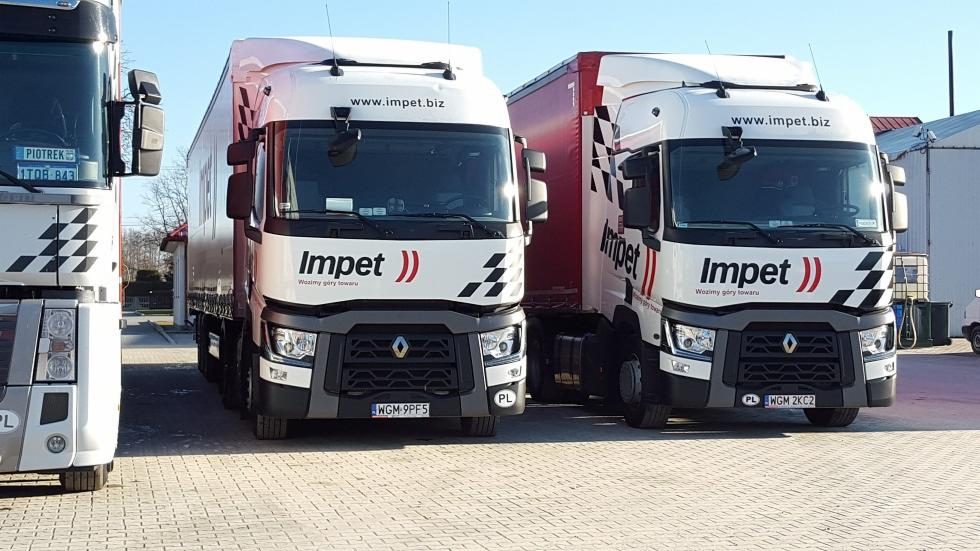 Dwa białe samochody ciężarowe z naczepami i logo firmy 'Impet', zaparkowane obok siebie na placu, widoczne tablice rejestracyjne i hasło 'Wozimy góry towaru'.