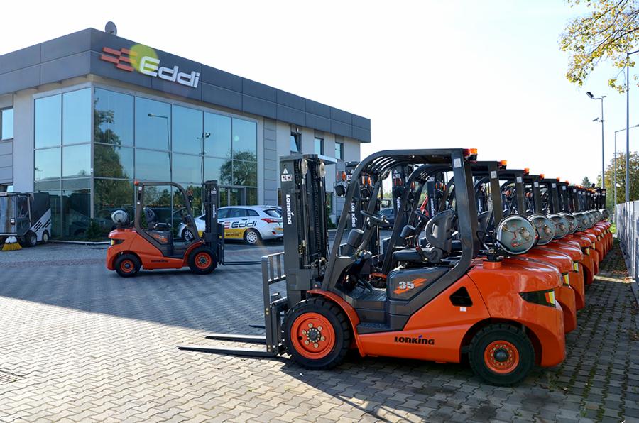 Rząd nowych, pomarańczowych wózków widłowych marki Lonking, ustawionych przed budynkiem firmy Eddi z dużymi oknami i logo na dachu.