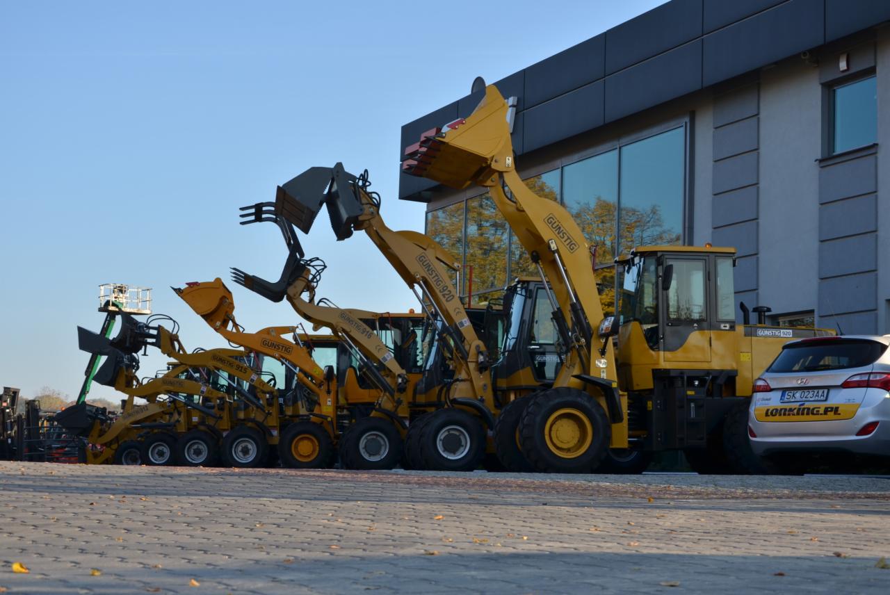 Rząd nowych, żółtych wózków widłowych marki Gunstig, o różnych modelach, ustawionych na placu przed budynkiem z dużymi oknami, z widocznym samochodem osobowym w tle.