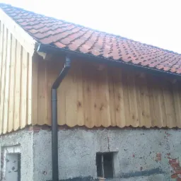 Wykończona drewnem szczytowa ściana budynku z czerwoną dachówką i czarną rynną. Ściana betonowa z ceglanymi elementami. Widok z dołu.