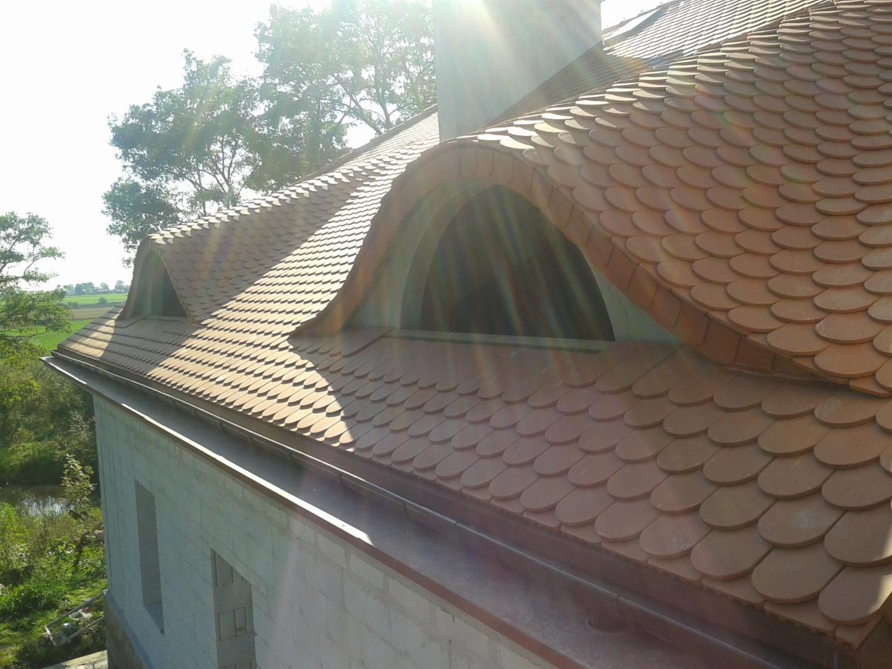 Dach pokryty łuską ceramiczną z oknami połaciowymi w kształcie łuków, na tle zieleni i błękitnego nieba z promieniami słońca. Rynna w kolorze dachu.