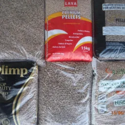 Różne marki pelletu drzewnego w przezroczystych workach, widoczne etykiety: Lava Premium Pellets, Olimp Top Quality Pellets, Barlinek Wooden Pellets. Jeden worek z datą produkcji 13/06/2016.