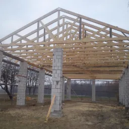 Konstrukcja drewniana dachu w trakcie budowy, wsparta na murowanych filarach z bloczk&oacute;w, widok na zewnątrz w pochmurny dzień.