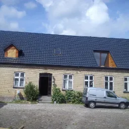 Duży, jasny budynek z ciemnym dachem z dachówki, widoczne okna dachowe i fragment okna w szczycie. Na podwórku zaparkowany srebrny van.