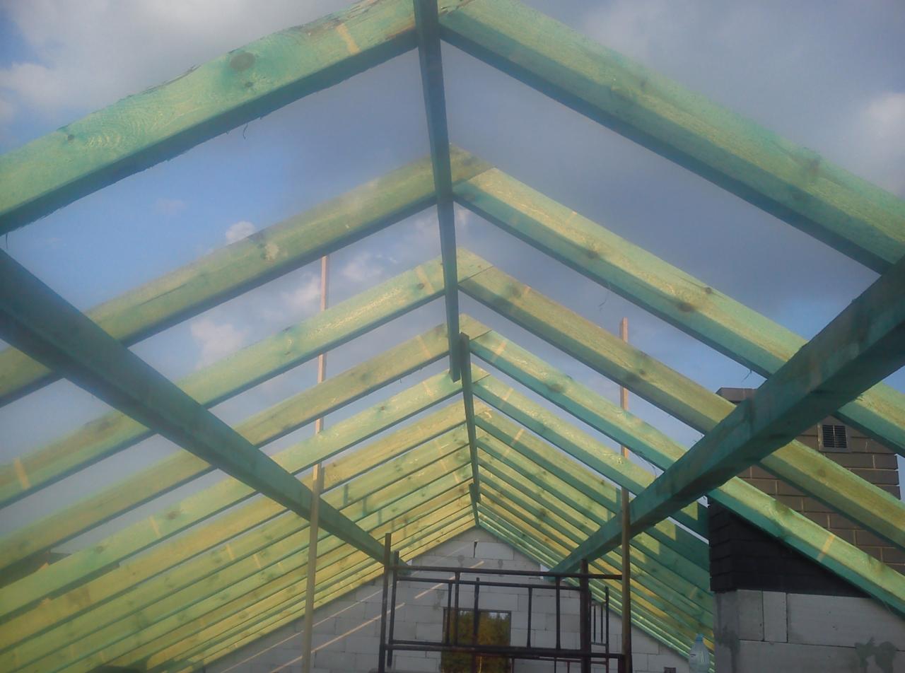 Konstrukcja drewniana dachu z impregnacją w kolorze zielonym, widok od wewnątrz, na tle nieba. Widoczne elementy murowane budynku.