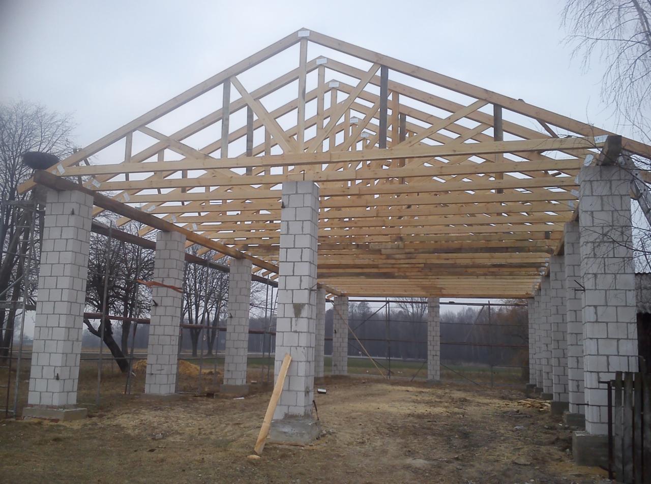 Konstrukcja drewniana dachu w trakcie budowy, wsparta na murowanych filarach z bloczków, widok na zewnątrz w pochmurny dzień.