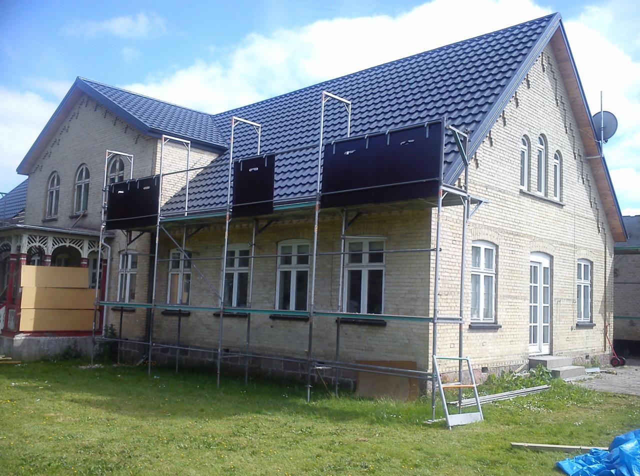 Dom z jasnej cegły z szarym dachem, częściowo osłonięty rusztowaniem z czarnymi panelami. Widoczny fragment trawnika i błękitne niebo z chmurami.