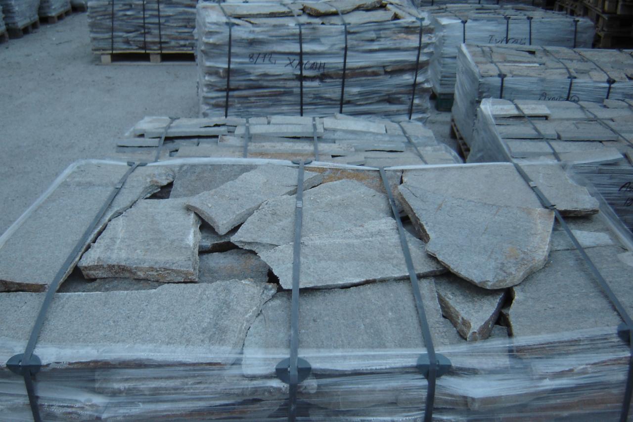 Palety z nieregularnymi płytami kamiennymi w szarym odcieniu, zabezpieczone folią i taśmami, składowane na zewnątrz.