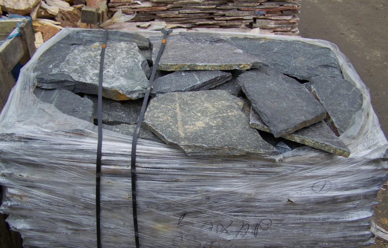 Paleta z nieregularnymi płytami kamiennymi w odcieniach szarości, zabezpieczona folią i czarnymi taśmami, widoczne inne palety z kamieniem w tle.