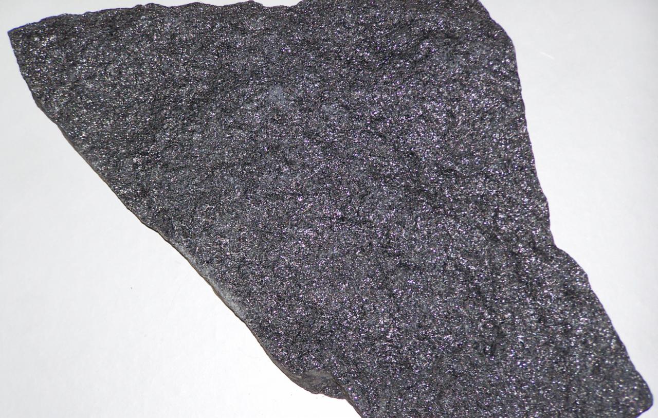 Szorstki, ciemnoszary element betonowy o nieregularnym kształcie, sfotografowany na jasnym tle, ukazujący jego chropowatą strukturę i drobne, błyszczące inkluzje.