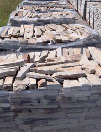 Palety z nieregularnymi płytkami kamiennymi w odcieniach beżu i szarości, zabezpieczone folią stretch, składowane na zewnątrz.