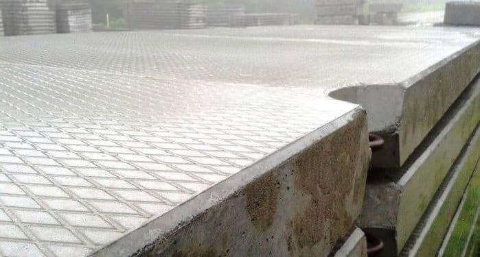 Stos betonowych płyt chodnikowych z wyraźnym wzorem rombów, widoczne stalowe uchwyty transportowe, ułożone na zewnątrz w pochmurny dzień.