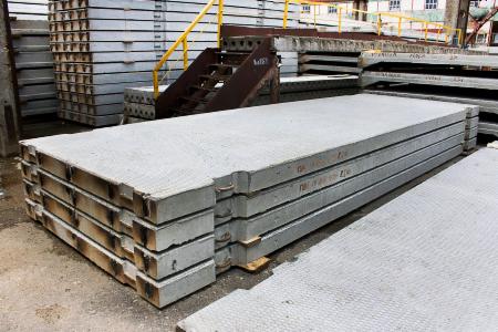 Stos płyt betonowych z widocznymi oznaczeniami transportowymi, składowane na placu budowy, w tle schody z żółtą barierką.