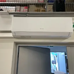 Biały klimatyzator marki Gree zamontowany na ścianie w pomieszczeniu magazynowym, widoczny znak ewakuacyjny i regały z towarem.