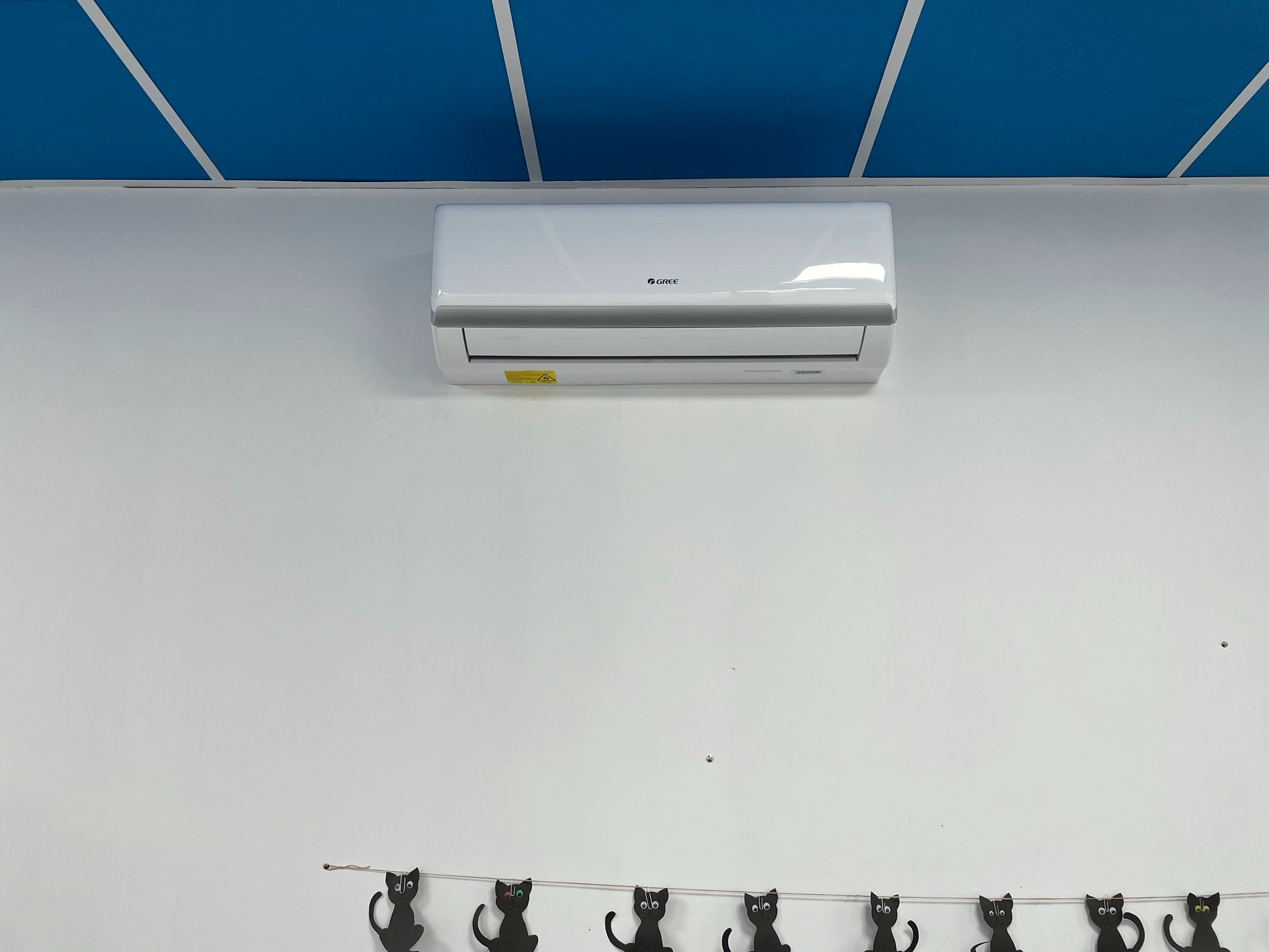 Biały klimatyzator marki Gree zamontowany na białej ścianie pod niebieskim sufitem z białymi pasami, poniżej dekoracja z czarnych kotów na sznurku.