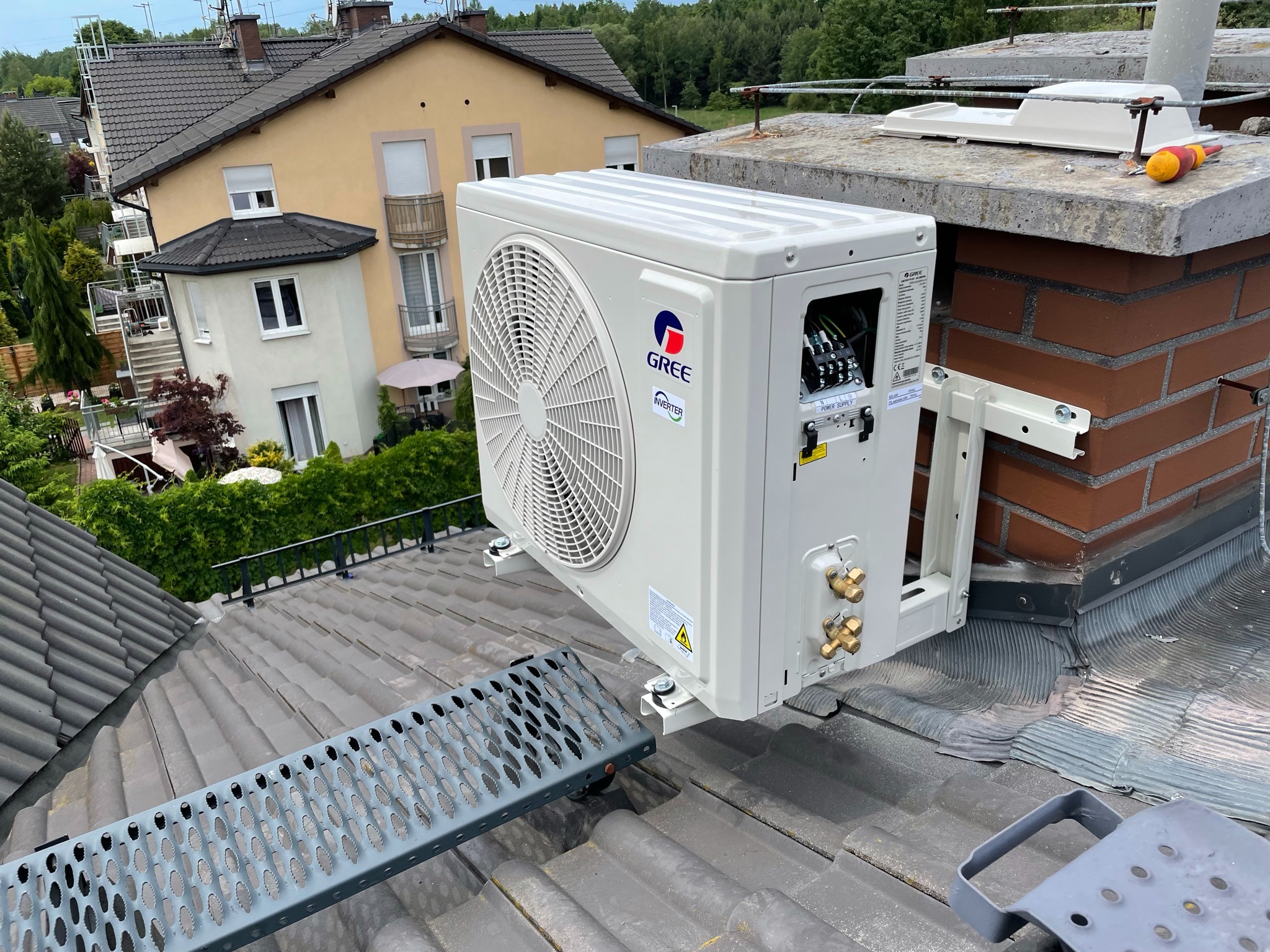 Jednostka zewnętrzna klimatyzacji GREE zamontowana na dachu budynku jednorodzinnego obok komina, widoczna drabina dachowa i narzędzia.