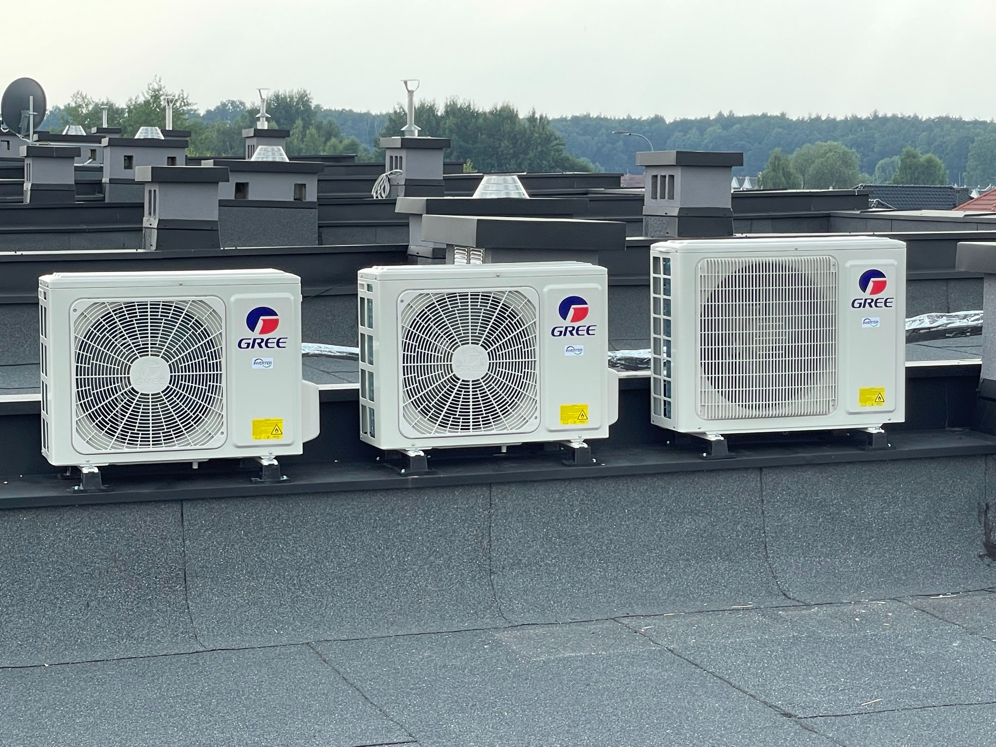 Trzy białe jednostki zewnętrzne klimatyzacji marki Gree, zamontowane na płaskim, czarnym dachu budynku z widocznymi kominami i drzewami w tle.