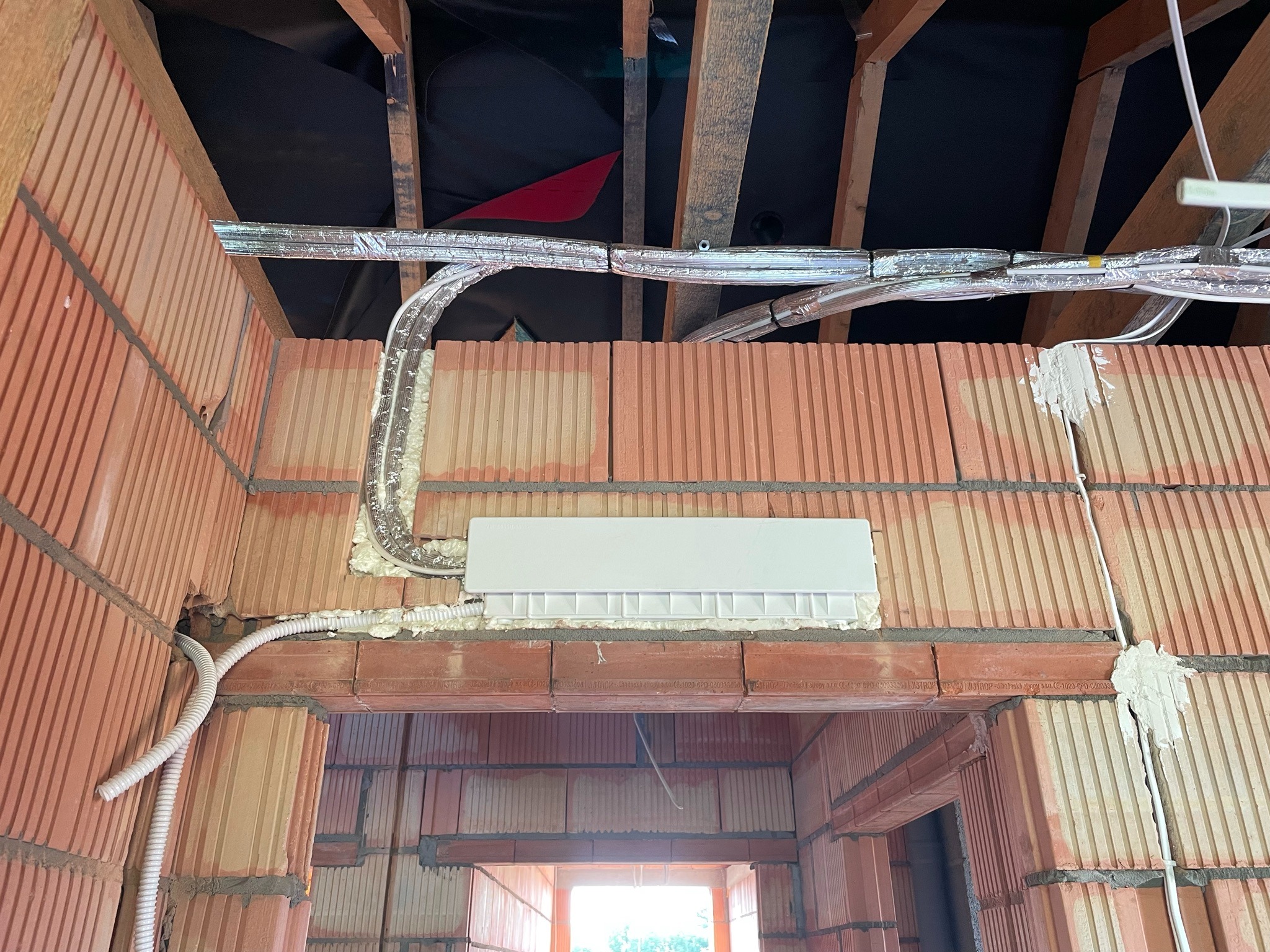 Montaż instalacji elektrycznej i klimatyzacji w surowym stanie budynku z cegły, widoczne kable w peszlach, rury w izolacji aluminiowej oraz biała obudowa urządzenia zamontowana w ścianie z pianką...